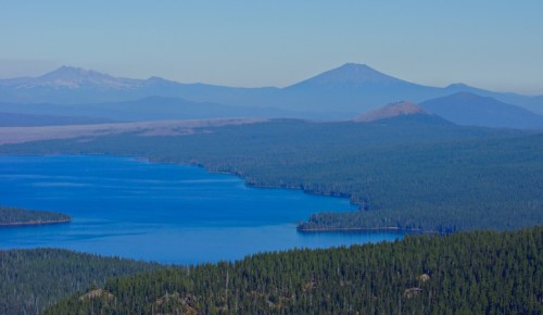 A panoramic view of a blue lake surrounded by dense forests and distant mountains under a clear sky.