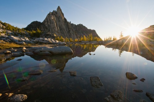 A stunning mountain peak reflected in a calm lake at sunset, surrounded by trees and rocky terrain.