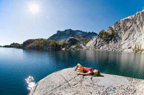 A person relaxes on a rock by a serene lake, surrounded by mountains and clear blue skies. Sunlight sparkles on the water.