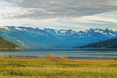 A serene lake surrounded by mountains, with grassy shores and a cloudy sky reflecting on the water's surface.