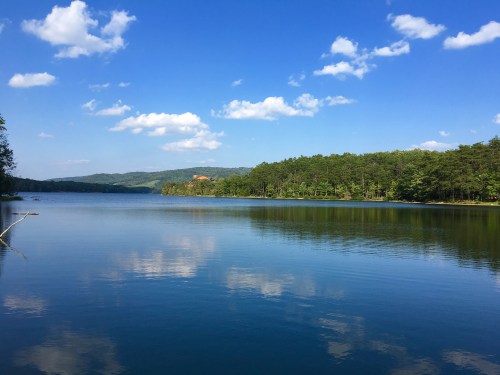 A serene lake surrounded by lush green trees under a blue sky with fluffy clouds reflecting on the water's surface.