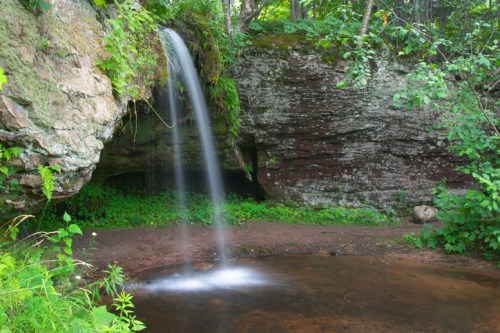 A serene waterfall cascades over a rocky ledge into a calm pool, surrounded by lush greenery and trees.