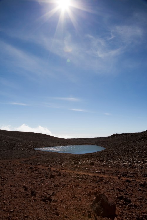 A sunlit landscape with a small pond surrounded by rocky terrain under a clear blue sky.