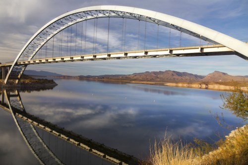 A modern arch bridge spans a calm lake, reflecting the sky and surrounding mountains in the water below.