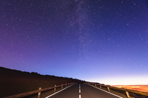 A winding road under a starry night sky, with a gradient of colors at the horizon.