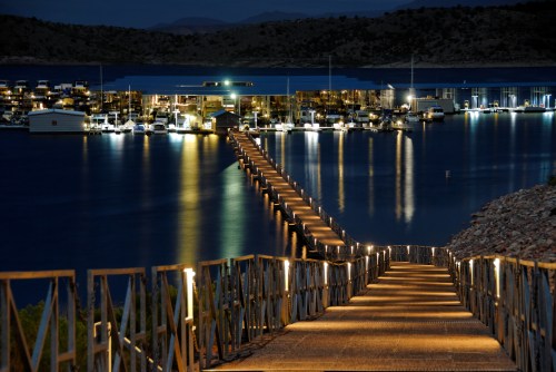 A lit walkway leads to a marina at night, reflecting lights on calm water with boats docked in the background.