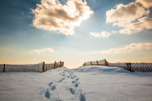 A snowy landscape with footprints leading to a wooden fence under a bright sky with fluffy clouds.