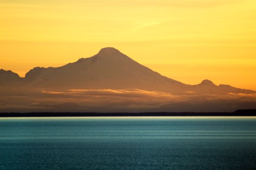 A majestic mountain silhouette against a vibrant orange and yellow sunset, reflecting on calm waters below.
