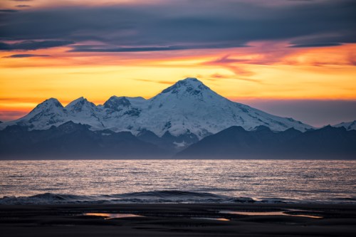 A stunning sunset over snow-capped mountains, reflecting on calm ocean waters with a colorful sky.