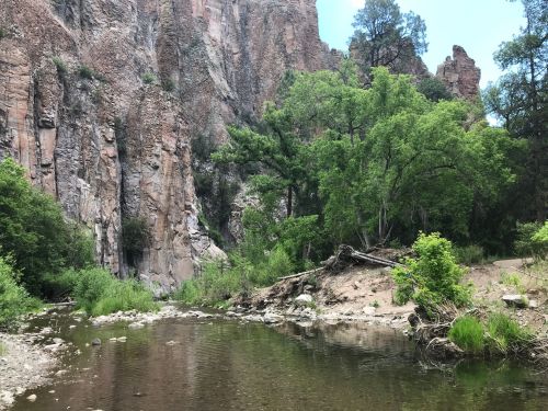 A serene river flows through a rocky canyon, surrounded by lush greenery and towering cliffs under a clear blue sky.