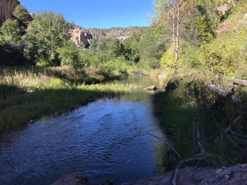 A serene river flows through a lush green landscape, surrounded by trees and rocky cliffs under a clear blue sky.