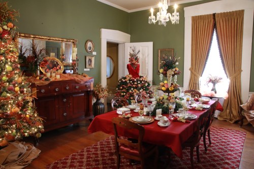A festive dining room with a red table set for a meal, decorated Christmas trees, and elegant holiday decor.