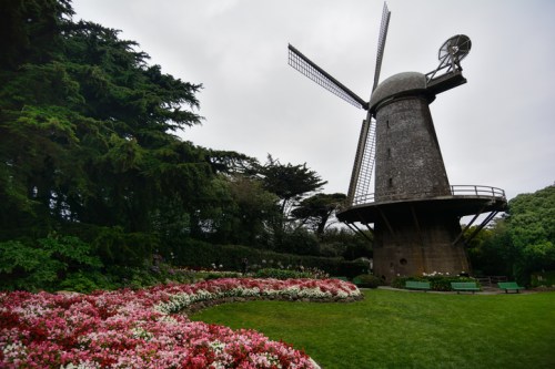 A windmill stands amidst lush greenery and colorful flower beds in a serene garden setting.