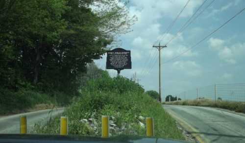 A roadside historical marker stands beside a rural road, surrounded by greenery and under a partly cloudy sky.