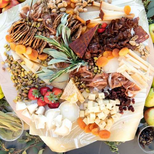 A colorful charcuterie board featuring cheeses, meats, nuts, fruits, and herbs arranged artfully.