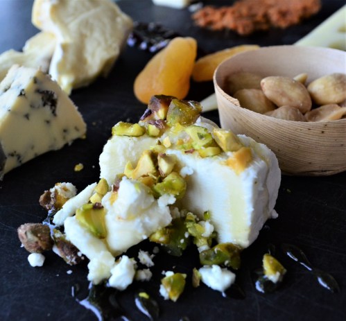 A close-up of a cheese platter featuring various cheeses, nuts, dried fruits, and a cheese topped with pistachios.