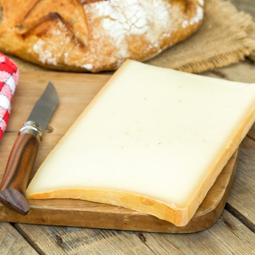 A block of cheese on a wooden cutting board with a knife and a loaf of bread in the background.
