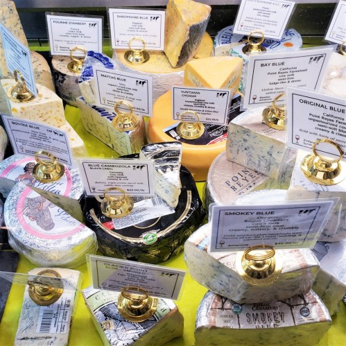 A variety of cheeses displayed in a case, each labeled with names and descriptions on small signs.