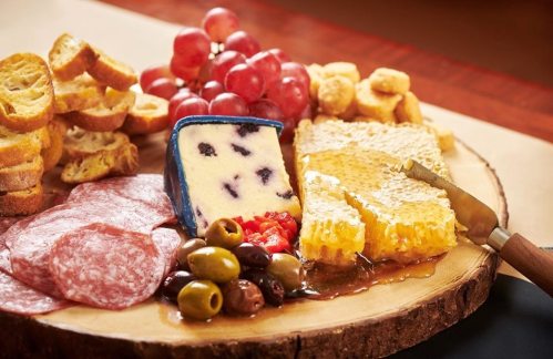 A wooden platter featuring assorted cheeses, salami, olives, grapes, honeycomb, and toasted bread slices.