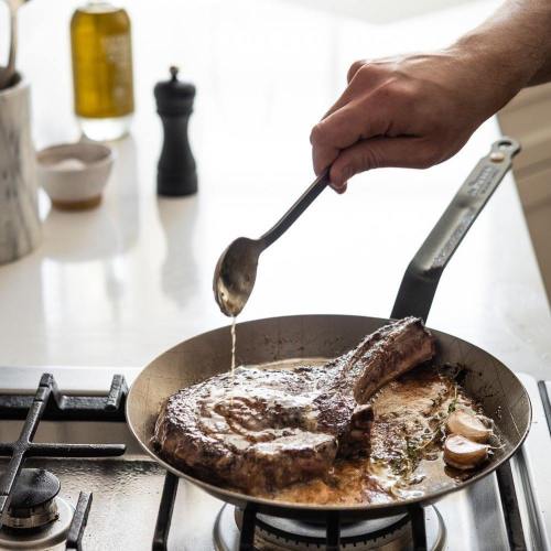 A hand drizzles sauce over a sizzling steak in a frying pan on a stovetop, with olive oil and spices in the background.