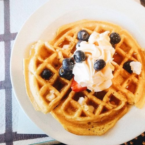 A plate of waffles topped with whipped cream, blueberries, and a strawberry, drizzled with syrup.