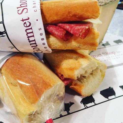 A close-up of a sandwich with salami in a baguette, alongside a block of cheese and a wrapped roll on a patterned surface.
