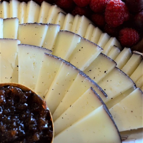A circular platter of sliced cheese, accompanied by a bowl of dark jam and fresh raspberries.