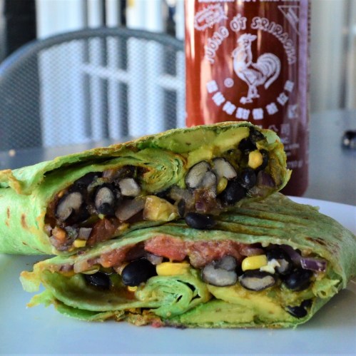 A cut green wrap filled with black beans, corn, and tomatoes, served with a bottle of Sriracha sauce.