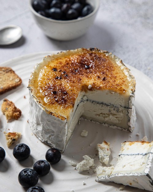 A round cheese with a caramelized top, partially sliced, surrounded by blueberries and crackers on a white plate.