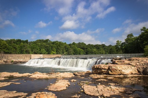 A serene waterfall cascades over a rocky ledge, surrounded by lush greenery and a clear blue sky.