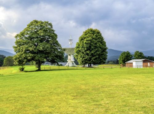 A grassy field with two large trees, a satellite dish, and a small building under a cloudy sky.