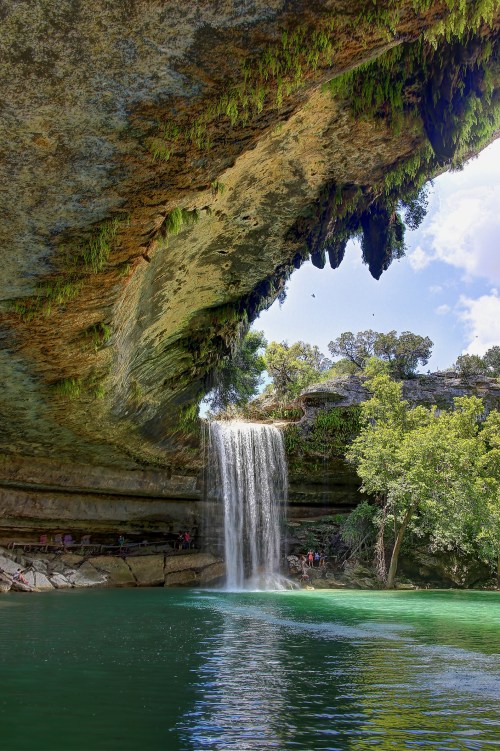 A serene waterfall cascades from a rocky overhang into a clear green pool, surrounded by lush trees and a bright sky.