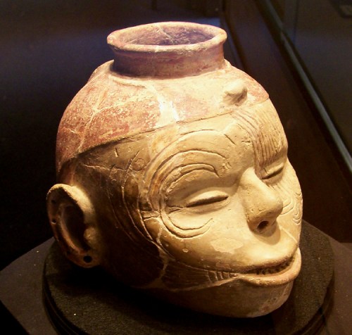 Ceramic head sculpture with intricate facial features and a small vessel on top, displayed in a glass case.