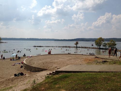 A sandy beach with people swimming and relaxing by a lake under a partly cloudy sky. Trees line the shore in the background.