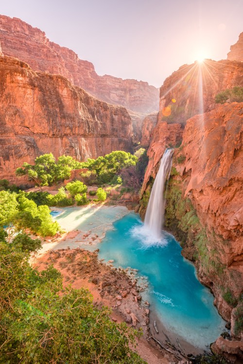A stunning waterfall cascades into a turquoise pool, surrounded by red rock cliffs and lush greenery at sunset.