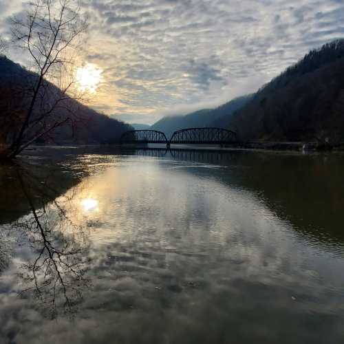 A tranquil river scene at sunset, featuring a bridge and reflections on the water, surrounded by hills and trees.