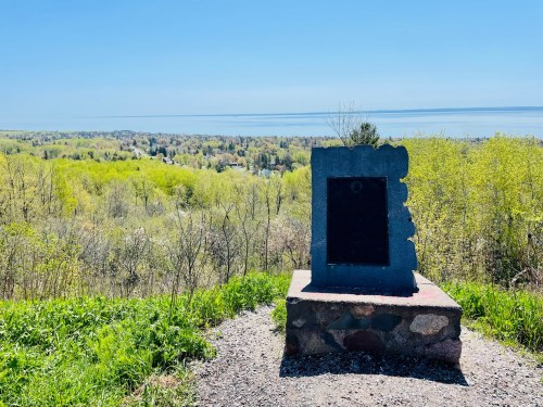 A stone monument stands on a hill overlooking a lush green landscape and a calm body of water under a clear blue sky.