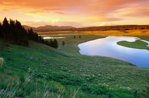 A serene landscape featuring a winding river, lush green fields, and a colorful sunset over distant mountains.