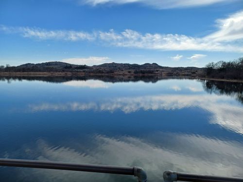 A serene lake reflecting blue skies and clouds, surrounded by hills and trees under a clear day.