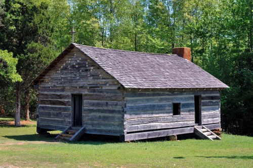A rustic wooden cabin with a sloped roof, set in a green landscape surrounded by trees.