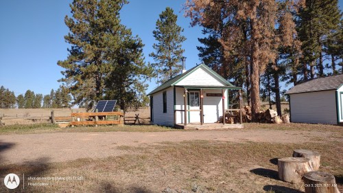 A small white cabin with a green roof, surrounded by trees and a solar panel, in a rural setting.