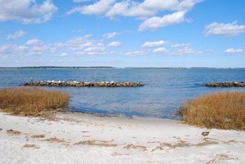A serene beach scene with sandy shore, calm water, and a rocky jetty under a blue sky with fluffy clouds.