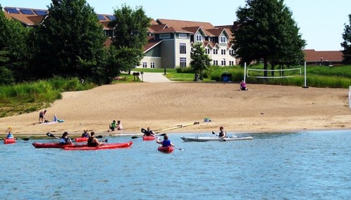 A scenic view of kayakers on a lake near a sandy beach and a large building in the background, surrounded by trees.