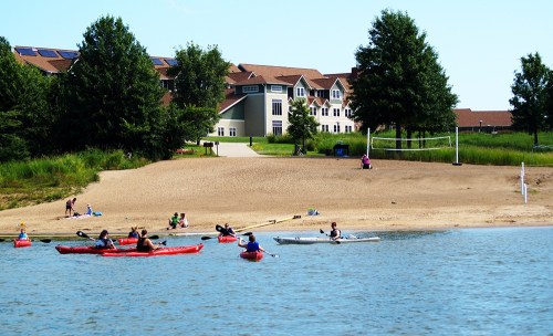 A sunny beach scene with kayakers on the water and a sandy shore with a volleyball net and a building in the background.