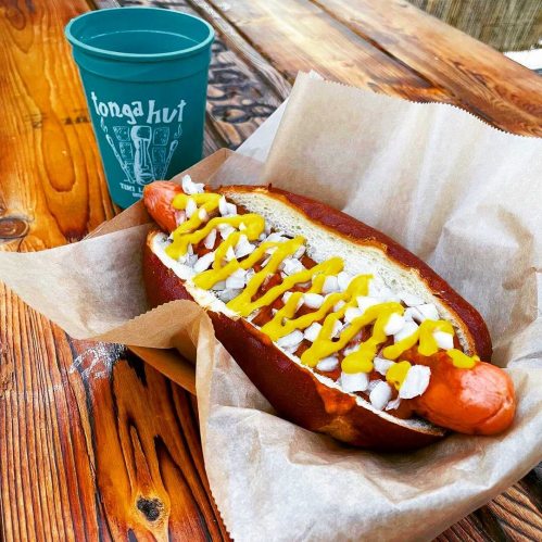 A hot dog in a pretzel bun topped with mustard and onions, served with a blue cup on a wooden table.