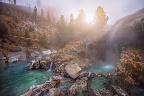 A misty landscape featuring a serene river, rocky terrain, and cascading waterfalls under a soft, glowing sun.