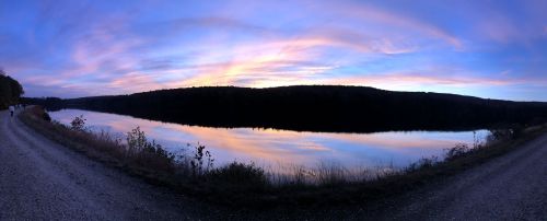 A serene lake at sunset, reflecting colorful clouds and surrounded by trees, with a gravel path along the shore.