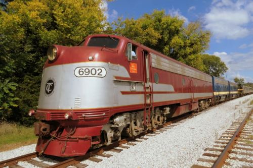 A vintage red and silver train engine on a gravel track, surrounded by green trees and a blue sky.