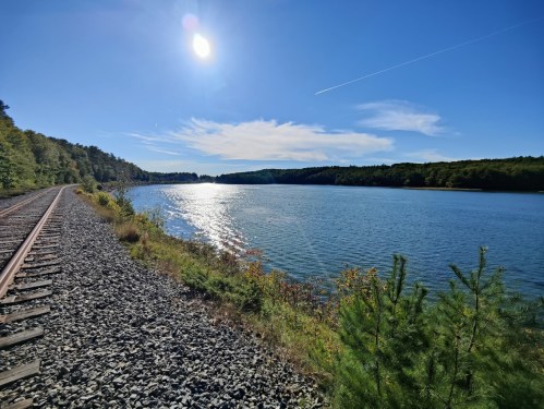 A serene lake reflects sunlight, with a railway track running alongside and trees lining the shore under a clear blue sky.