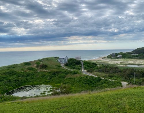 A coastal landscape featuring green hills, a pond, and a cloudy sky over the ocean in the distance.